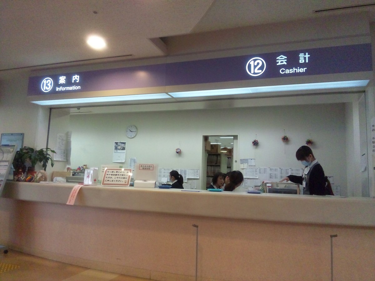 A long counter in a Japanese hospital lobby beneath a purple sign band reading Information on the left and Cashier on the right, the back wall papered with white notices, a masked visitor standing at the cashier window