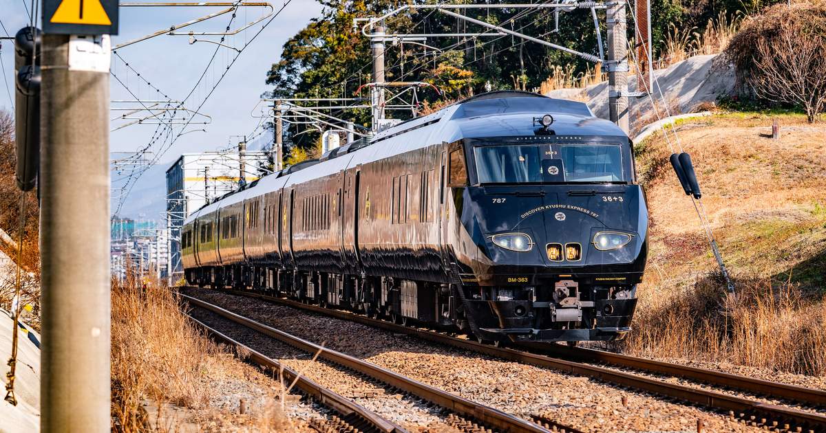 全身漆黑的 36ぷらす3 從彎道迎面開來,車頭擋風玻璃下方印著金色的 DISCOVER KYUSHU EXPRESS 787 與 36+3,兩側是冬天枯黃的草坡和電線桿