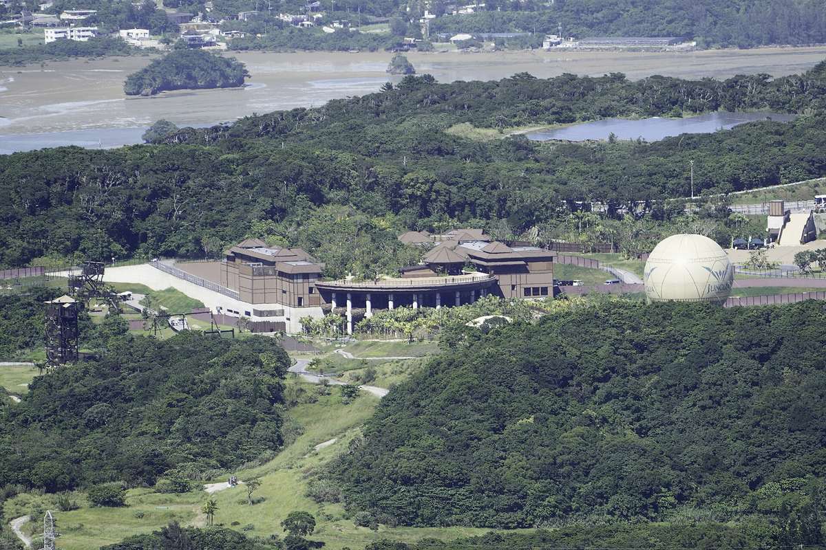 The JUNGLIA site in northern Okinawa seen from a hilltop: brown pitched-roof buildings and a white colonnade enclosed by unbroken subtropical broadleaf forest, a cream-coloured balloon lettered JUNGLIA standing to the right, a steel viewing tower to the left, and a tidal inland sea with a settlement beyond