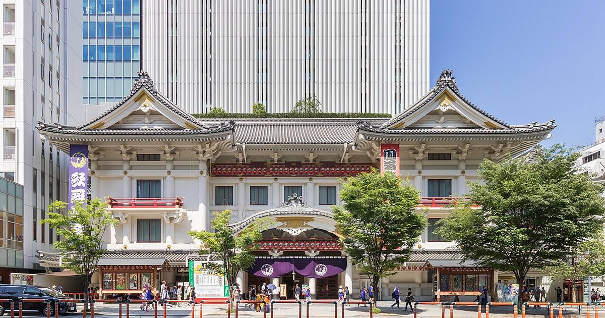 The full front elevation of the Kabukiza theatre in Ginza on a clear day: two white gabled roofs flanking a curved karahafu canopy over the entrance, a row of red lanterns beneath the eaves, a purple curtain bearing the phoenix crest at the doorway, and the Kabukiza Tower rising behind