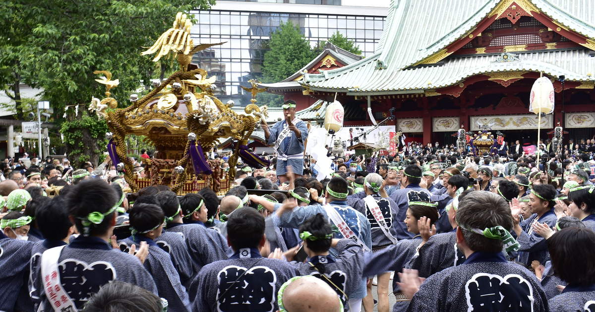 A gilded portable shrine carried into the Kanda Myojin precinct during Kanda Matsuri, bearers in matching indigo happi coats and green-and-white headbands, the vermilion main hall and its green copper roof behind