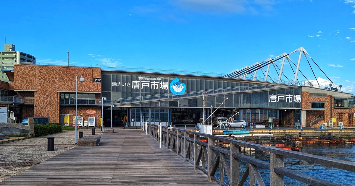下關唐戸市場的正面外觀:海邊木棧道通往市場,玻璃立面上寫著「活きいき 唐戸市場」和藍色河豚標誌,右側是白色鋼架屋頂,晴天藍天