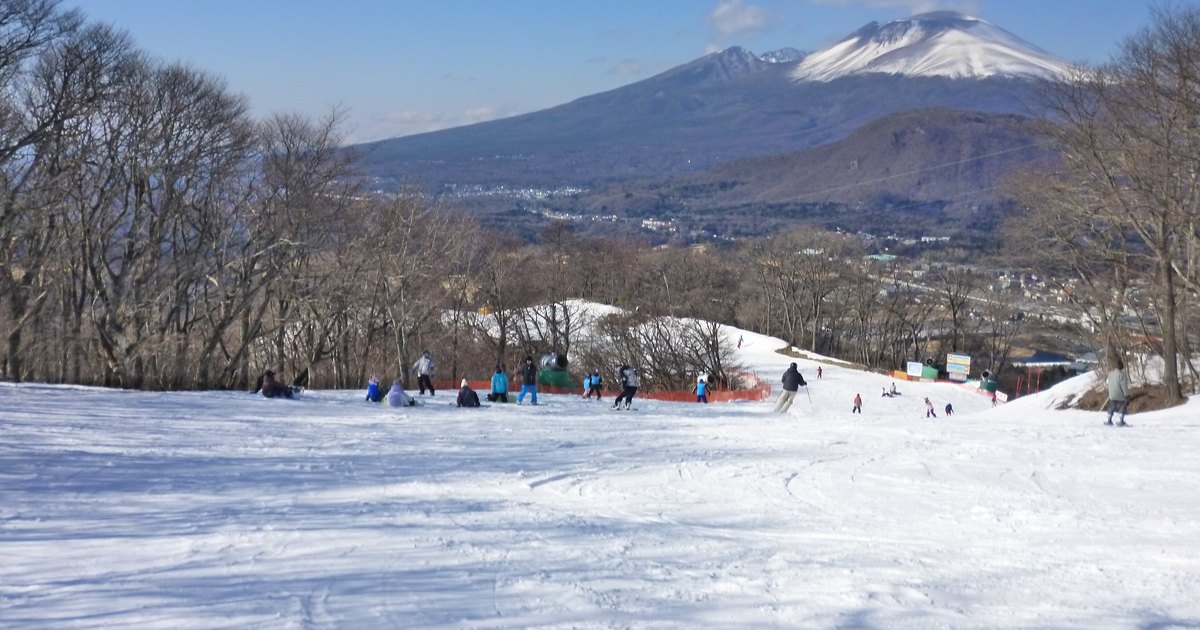 輕井澤王子滑雪場的雪道，滑雪客坐在坡邊休息，兩旁是落葉的樹林，正前方是山頂積雪的淺間山（2012 年 1 月攝）