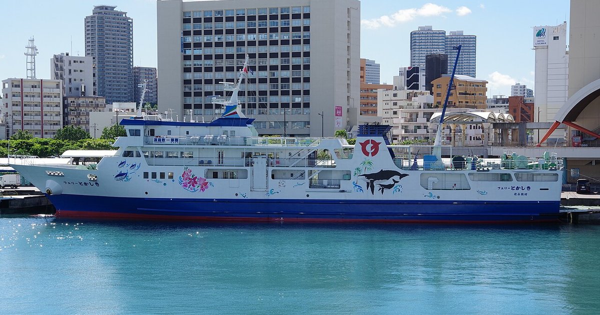 The white and blue Ferry Tokashiki berthed at Tomari Port in Naha, its hull painted with a large whale and a calf alongside tropical fish and hibiscus flowers, Naha high-rises behind