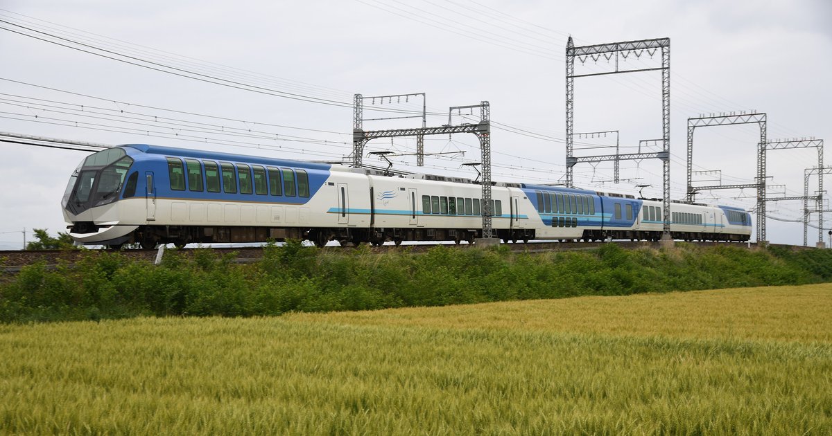 近鐵觀光特急島風號從側面駛過:六節白色車廂,頭尾兩節的上半部是藍色、開著大片車窗,前景是一片金黃的麥田
