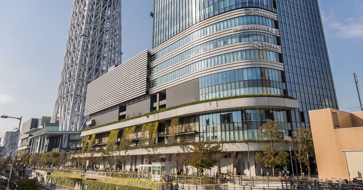 晴天下的東京晴空街道 East Yard：玻璃高樓底部是一圈圈弧形的商場樓層，外牆掛著 Tokyo Solamachi 的標誌，左邊是晴空塔白色鋼骨的塔腳