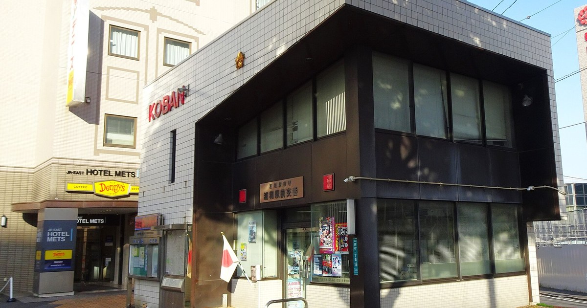 A koban outside Urawa Station in Japan: red three-dimensional KOBAN lettering on a white-tiled corner building, a bronze nameplate and gold police emblem under the dark eaves, a Japanese flag beside the door, wanted and public-notice posters covering the glass entrance, with JR-EAST HOTEL METS and Denny's signs next door