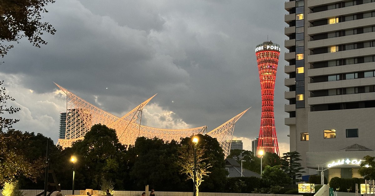 Meriken Park in Kobe at dusk: the white lattice roof of the Kobe Maritime Museum lit in warm light, Kobe Port Tower lit red, and Hotel Okura on the right; no Luminarie lights are in the picture
