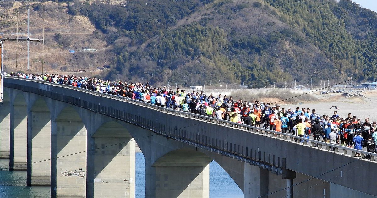 2018 年高知龍馬馬拉松,一長串穿彩色跑衣的跑者擠滿仁淀川河口大橋的橋面,從左往右跑,橋下是河口的水面與一排圓拱橋墩,背後是長滿樹的山與海邊沙灘