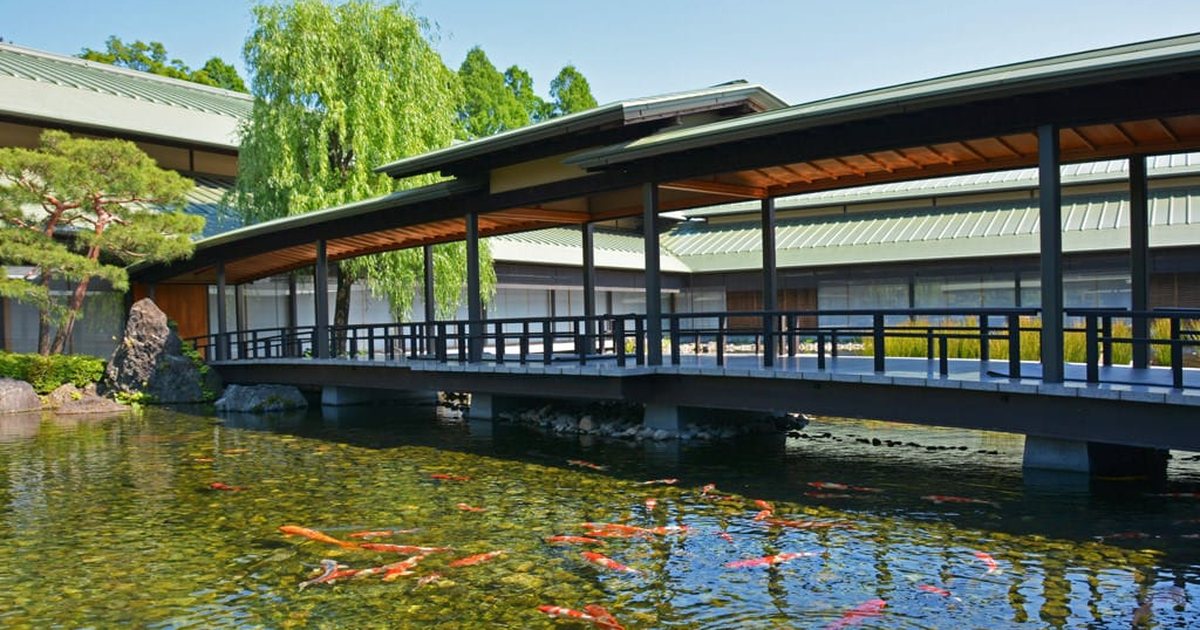 京都迎賓館庭園的廊橋：深色木欄杆、銅板屋頂的廊橋橫跨清澈的池塘，水裡游著許多錦鯉，後方是綠色屋頂的館舍與一棵垂柳