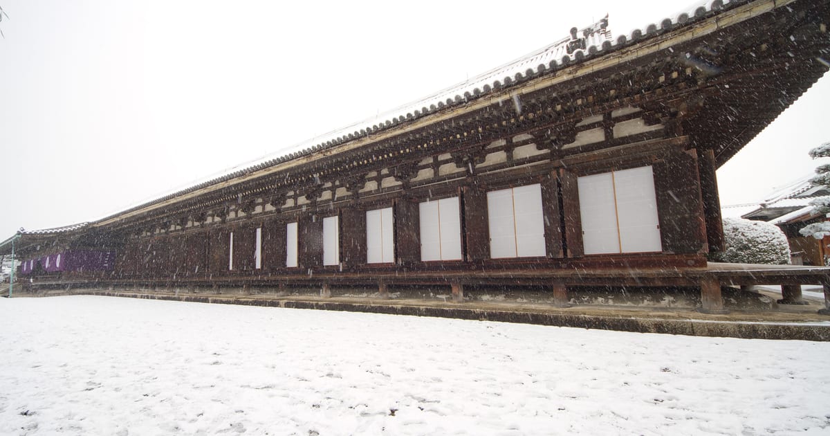 A long tiled temple roof in the Sanjusangendo quarter of eastern Kyoto running away into falling snow, its white shutter panels all closed, with an empty snow-covered forecourt in front marked only by a few footprints.