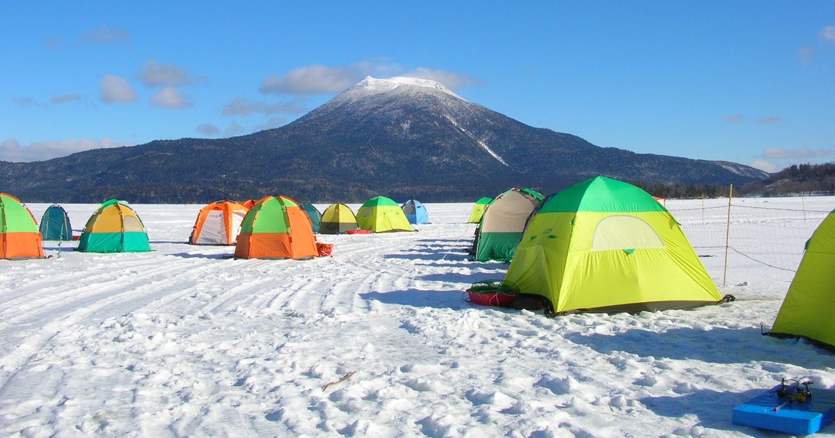 結冰積雪的阿寒湖湖面上散布著橘、綠、黃各色的西太公魚釣帳篷,雪地上有雪上摩托車壓出的車轍,遠方是山頂積雪的雄阿寒岳