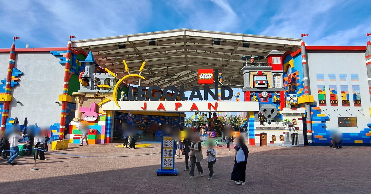 名古屋樂高樂園正門:白色大棚頂下掛著「LEGOLAND JAPAN」招牌與紅色 LEGO 標誌,兩側是積木造型的城堡與塔樓外牆,門前紅磚廣場上有排隊的遊客和一塊禁止攜帶物品的告示牌,藍天