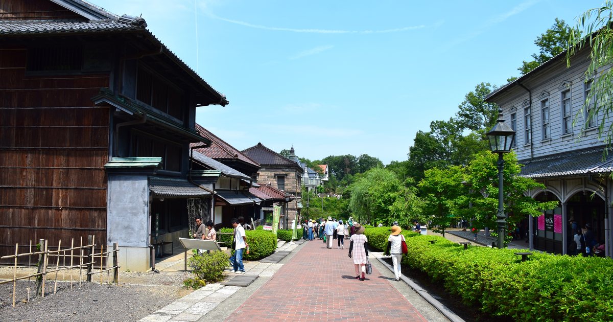 The red-brick street at Meiji-mura, dark timber Japanese buildings on the left, a white Western-style two-storey building on the right, a gas lamp between them and visitors walking away down the paving