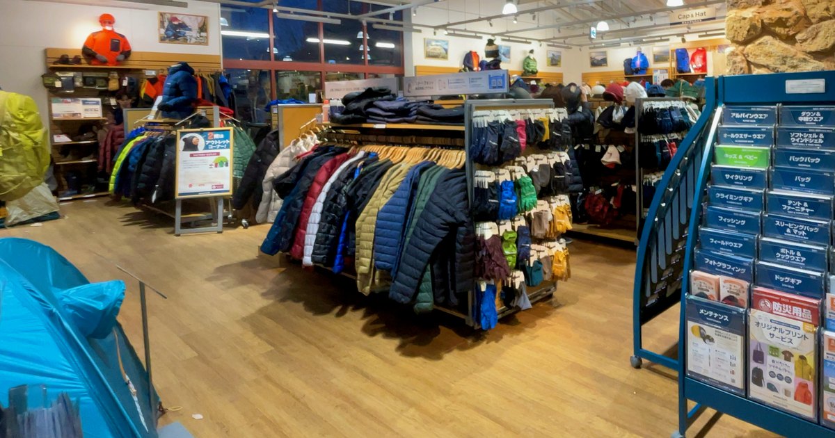 Montbell 門市店內:木地板中央一座掛滿深藍、紅、白、卡其色輕量羽絨外套的衣架,旁邊掛著成排手套,後方牆面是穿橘色雪衣的人形模特兒與帽子區,右側是放型錄的藍色架子,2023 年 12 月攝於愛知縣長久手店