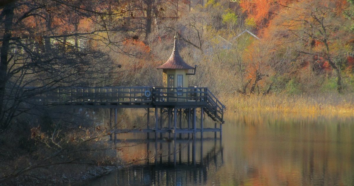 嚕嚕米樂園的宮沢湖畔:湖面上一座木頭棧橋,盡頭是黃牆、紅褐色尖頂的小木屋,背後山坡的樹轉成橘紅與黃色,湖面倒映著樹影,2021 年 12 月上旬