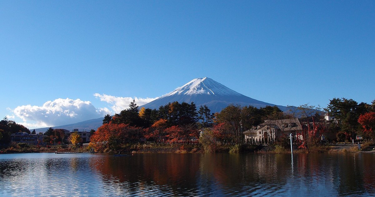 晴天早上的河口湖:只有山頂一截是白的富士山,山腰以下沒有雪,湖岸是轉紅轉黃的樹與幾棟房子,倒映在湖面上