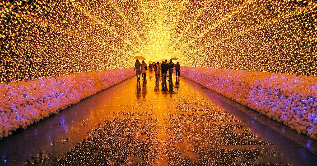 Inside the light tunnel at Nabana no Sato: warm gold LED bulbs cover the ceiling and both walls, a few visitors with umbrellas walk away from the camera, and the wet floor reflects the lights