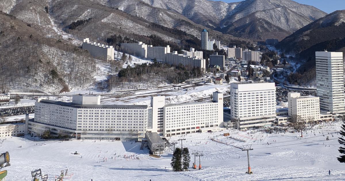 苗場滑雪場山腳:一整排白色的苗場王子飯店大樓沿著雪場底部排開,右側是一棟高塔樓,前景是壓雪雪道、吊椅纜車支柱和造雪機,後方是覆雪的山谷與成排公寓大樓