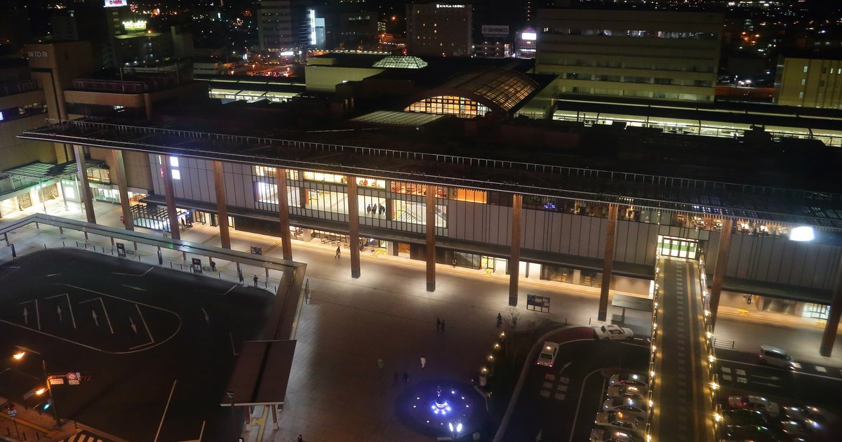 夜晚從高處俯瞰長野站善光寺口:整排木柱的車站正面亮著燈,左下是巴士圓環,站前廣場中央有一圈藍色燈光
