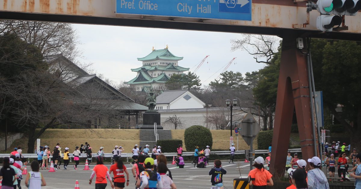 名古屋女子馬拉松比賽中的跑者背影跑過名古屋城東側的路口,遠方是綠色銅瓦屋頂的名古屋城天守與一座武將銅像,上方是標著縣廳、市役所的藍色路牌