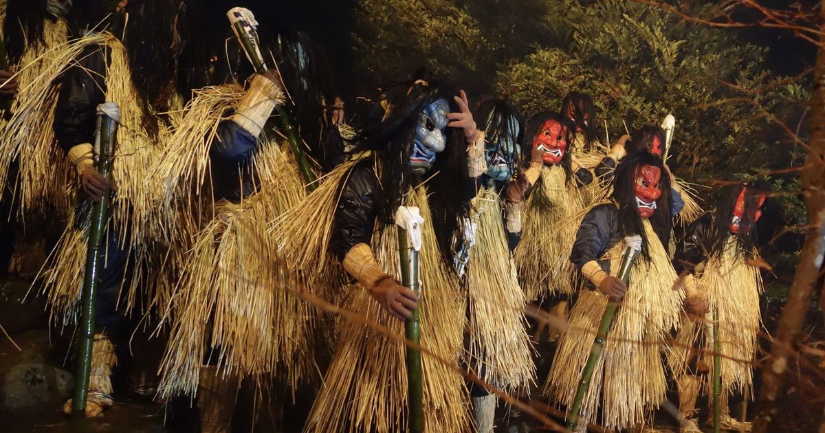 夜間的真山神社境內,一整排戴著面具的生剝鬼並排站著:由左至右是白面、藍面、紅面、褐面等各不相同的鬼面,全部披著及膝的稻草蓑衣、腰間繫著稻繩,每人手裡拄著一根綁了白紙的青竹棒,背景是黑暗中的常綠灌木與濕漉漉的石砌地面