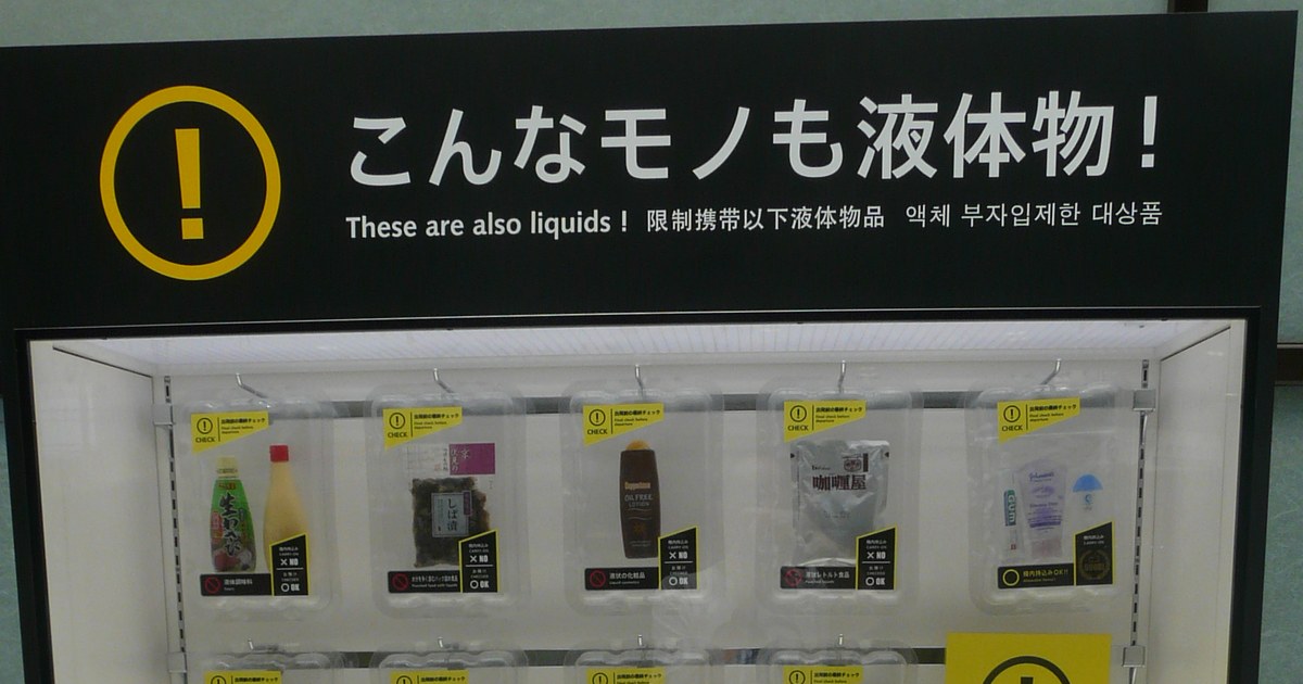 The liquids display case at Narita Airport: a black header panel with a yellow exclamation mark reads "These are also liquids!" in Japanese, English, Chinese and Korean, above a glass case whose top shelf holds five clear trays containing liquid seasoning, wet pickles, a bottle of lotion, a retort curry pouch, and a zip-lock bag of toothpaste and small tubes.