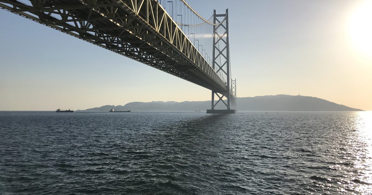 從神戶舞子海邊仰望明石海峽大橋,鋼桁架橋面從左上延伸到主塔,橋下海面對岸是傍晚逆光中的淡路島山稜