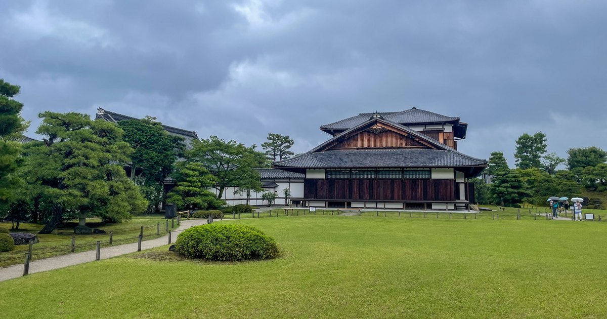 二條城本丸御殿外觀(2024 年 6 月攝,修理完工後):草坪後方是深褐色木板牆、灰瓦屋頂的御殿建築,後面還有一棟較高的樓閣,左側是松樹與通往御殿的砂石步道,天空多雲