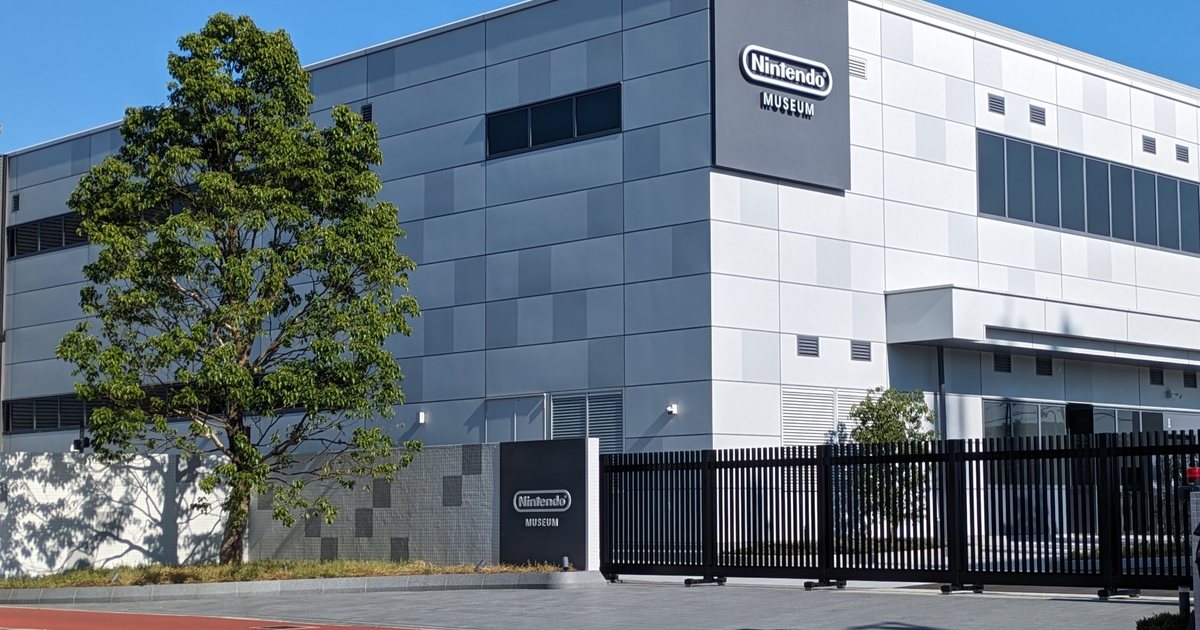 A museum exterior corner under a cloudless blue sky: the left half is clad in dark grey metal panels, the right half in white and pale grey panels, and a dark grey sign board on the corner carries the white oval Nintendo wordmark above the word MUSEUM; a leafy tree stands in front of the wall, a matching dark grey site monument bearing the same name sits on the low boundary wall, and a long black picket gate runs closed across the right of the frame above a grey paved footway and a red cycle lane