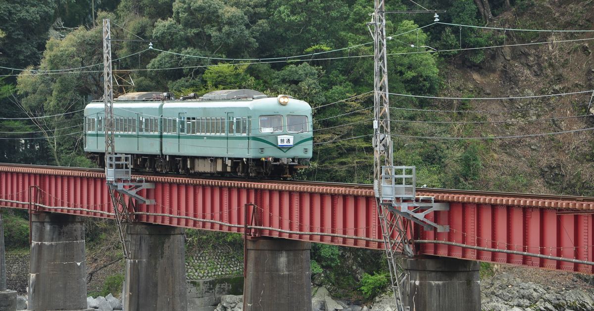 大井川鐵道的淺綠色普通電車開過紅色鐵橋，橋下是混凝土橋墩與青綠色的大井川，背景是山壁上的樹林