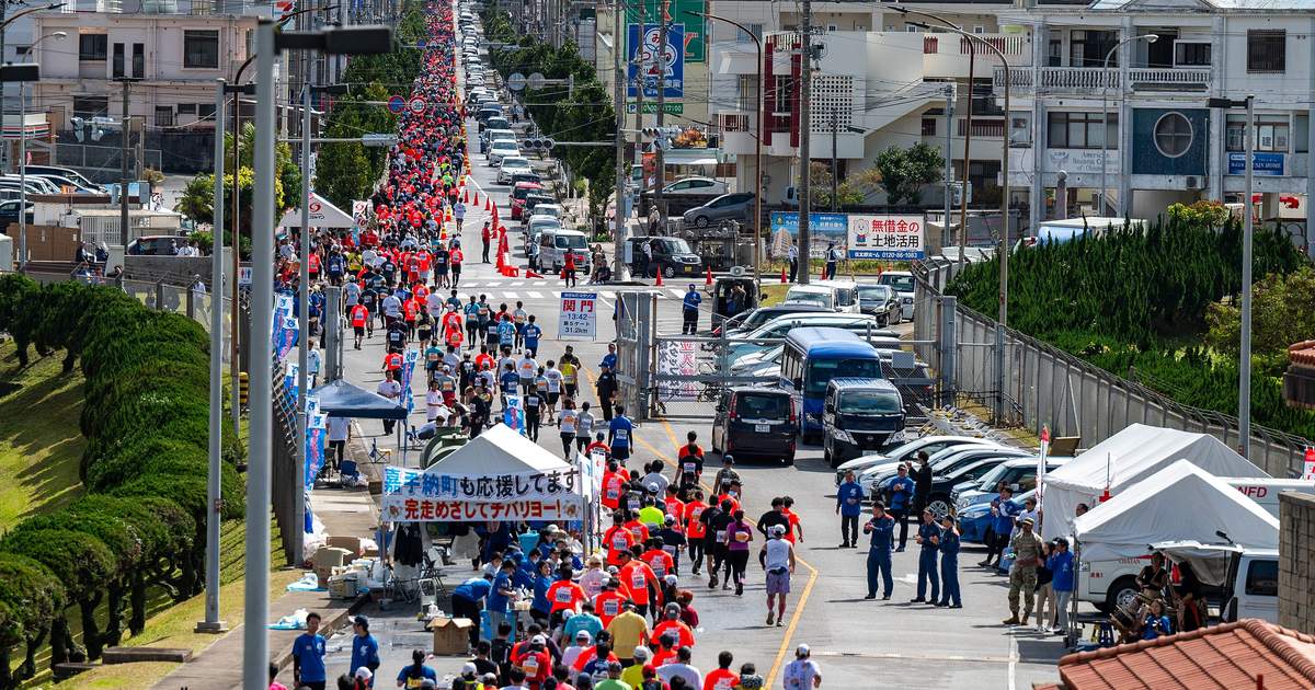 沖繩馬拉松 2025 年賽況:從高處往下看一條筆直的市區道路,上千名穿紅、白、藍衣服的跑者背對鏡頭往遠方跑,左側是修剪成圓球的綠籬與基地圍欄,路中央立著關門時刻牌,右側停著工作車輛與白色帳篷