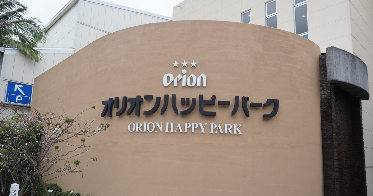 名護 Orion 啤酒工廠 Orion Happy Park 的入口招牌牆:一面米褐色弧形牆上嵌著三顆星與 Orion 標誌,下面是黑色大字「オリオンハッピーパーク」與白字 ORION HAPPY PARK;左邊有藍色停車場箭頭標示與一棵開著小白花的樹,牆後是白色廠房,陰天