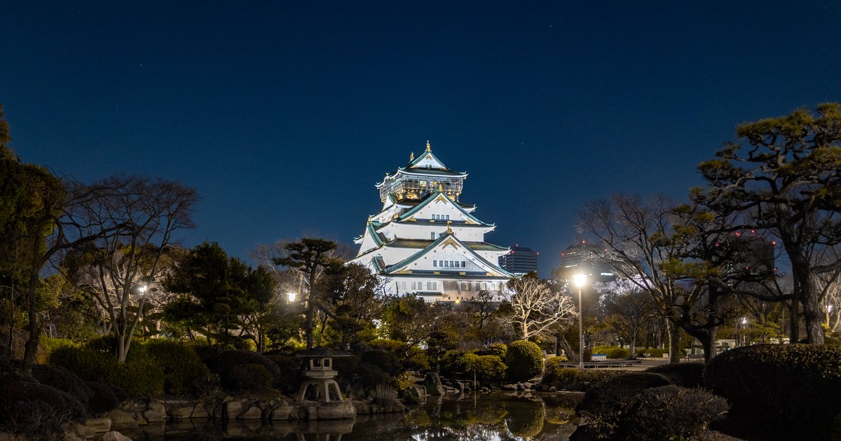2 月晚上的大阪城天守閣,白色外牆與綠色屋頂整棟打亮,前景是本丸日本庭園的水池、石燈籠和落葉的樹,池面映出天守閣的倒影