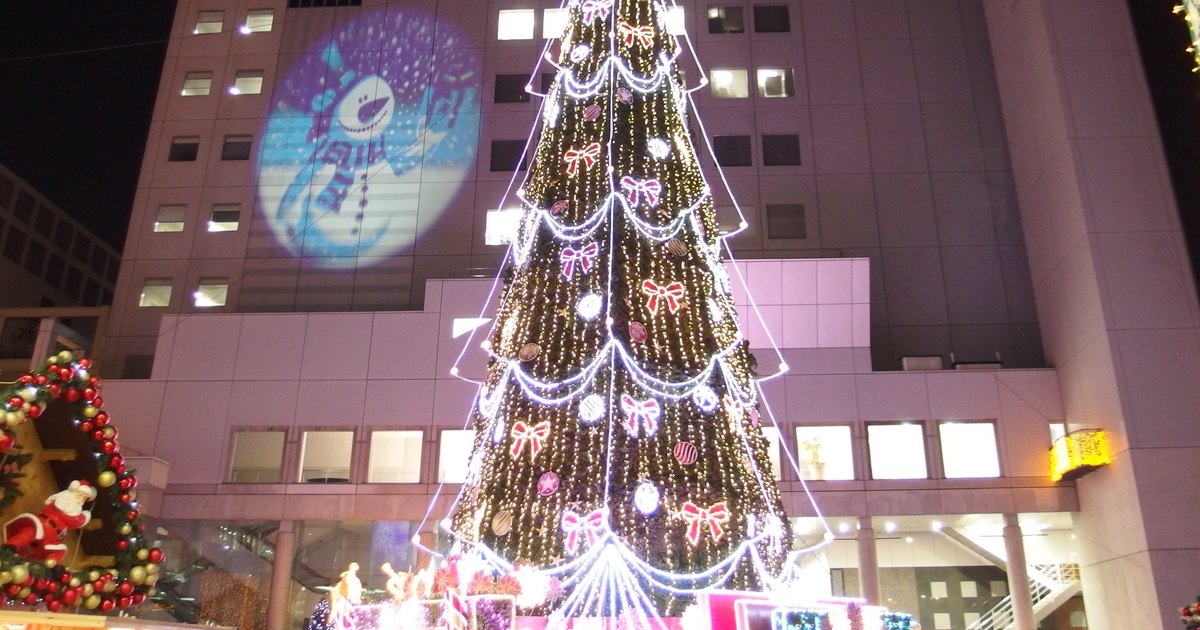 2014 年 12 月梅田藍天大廈 1 樓廣場的大型聖誕樹夜景:樹身掛滿白色燈串與紅白蝴蝶結燈飾,後方大樓牆面投影著雪人圖案,左下角是掛著紅金彩球花圈與聖誕老人偶的市集木屋屋頂