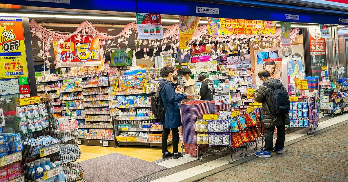The open frontage of a Japanese drugstore: three walls of shelving stacked from floor to ceiling with boxed medicines, toiletries and cosmetics under the arcade, pink cherry-blossom garlands and yellow sale banners hanging from the ceiling, a Tax-Free Shop sign on the right-hand pillar, and several masked shoppers browsing beside a stack of shopping baskets.