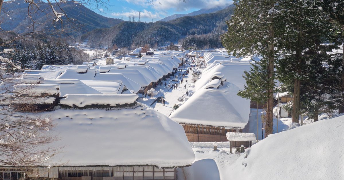 晴天從見晴台俯瞰冬天的大內宿：兩排茅葺屋頂蓋著厚雪，中間的老街上有零星遊客，遠方是積雪的山