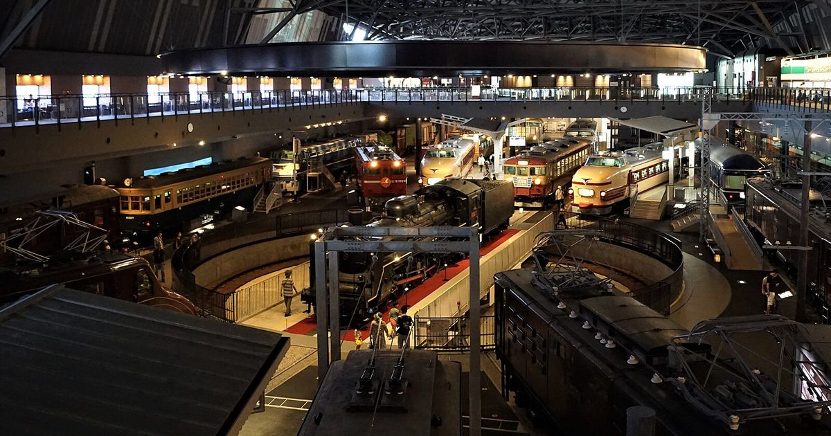 大宮鐵道博物館本館從二樓俯瞰車輛展示區:正中央圓形轉車台上停著黑色蒸汽機車 C57,四周呈放射狀排著紅色電力機車、奶油色紅帶的特急電車與藍色臥鋪客車,上方是鋼骨桁架屋頂與一圈二樓迴廊