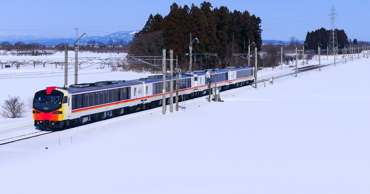晴天下一整片雪原，四節車廂的 Resort 白神號くまげら編成沿電氣化鐵道開過，車頭漆成黃、橘、紅三色，背景是一叢杉木林與遠方積雪的山