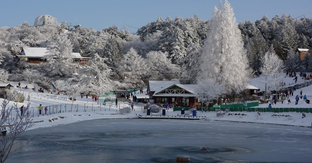 2014 年 2 月的六甲山スノーパーク：結冰的池塘後方是屋頂積雪的木造小屋與掛滿霧冰的樹林，右側緩坡上有遊客帶著雪盆玩雪，左上方山頂有一座圓頂建築