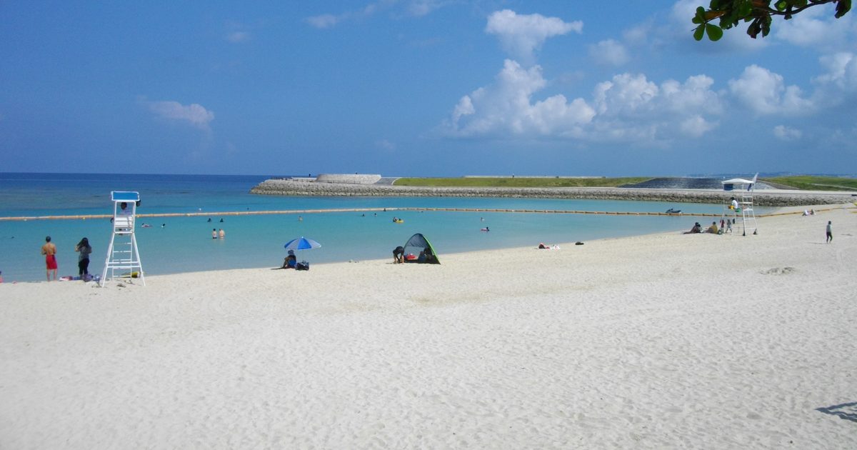 晴天白天的宜野灣トロピカルビーチ,白沙灘延伸到淺藍色的海,左邊立著白色救生員高台,遠處是石砌防波堤與草地,海面拉著一條浮球線