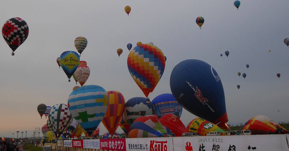 Dawn on the Kase River bank at the Saga Balloon Fiesta: dozens of hot air balloons, some already airborne and some still standing on the ground, behind a long line of sponsor banners