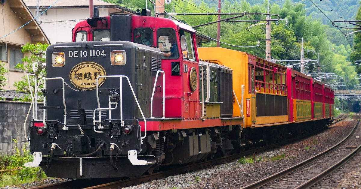 2026 年 7 月的嵯峨野小火車現行車：黑紅兩色的 DE10 1104 柴油機關車掛著「嵯峨野」圓牌，後面接一節黃色的 5 號車與四節紅色客車，行駛在嵐山一帶的綠色山腳