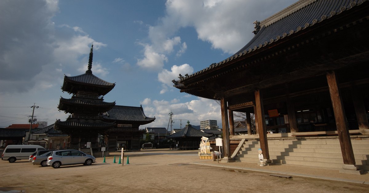 西大寺観音院的境內廣場：右側是本堂，深灰瓦頂與翹起的簷角、正面一排粗大的木柱與石階；左側遠處是三重塔，中間是一大片空曠的沙土地，幾輛車停在左邊，天空有大片積雲