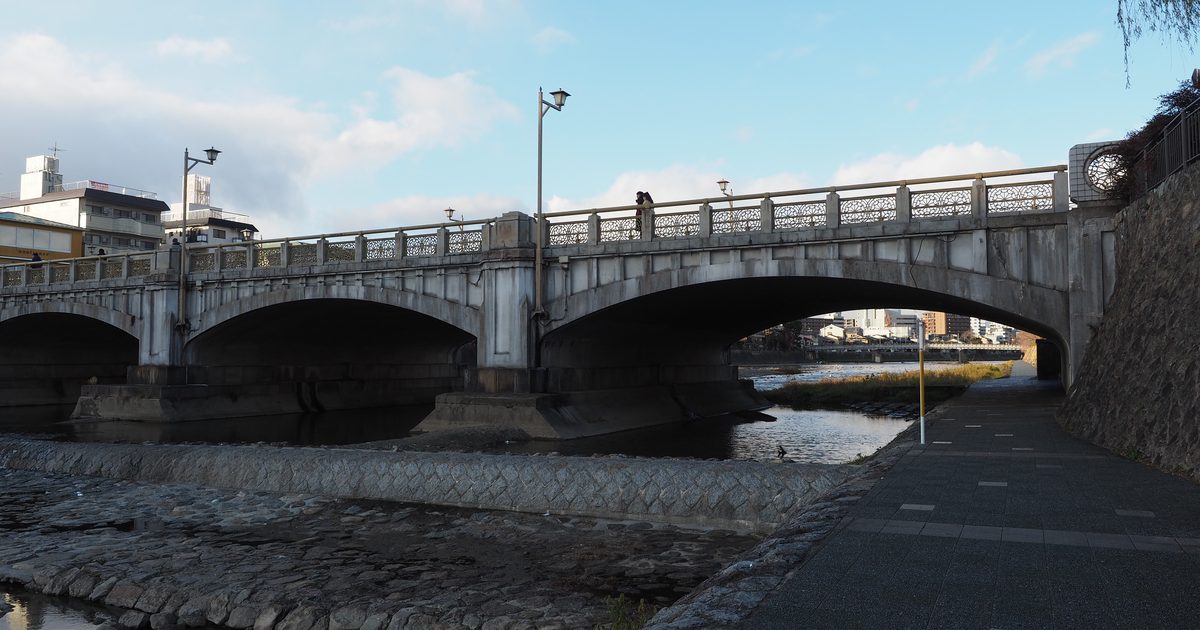 一月午後的七条大橋：從鴨川東岸河堤步道往上看，兩道寬闊的石造拱橋橫跨淺淺的河面，橋上欄杆是一格格鏤空的花紋，河床鋪著石塊，藍天裡飄著幾朵白雲