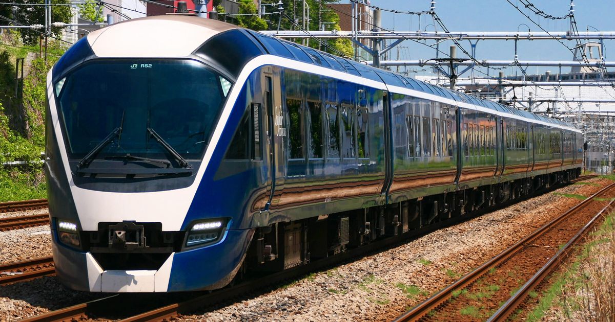 E261 系 SAPHIR 踴子號迎面駛來:深藍色車身、白色車頂與車鼻,擋風玻璃上方顯示 RS2,側面窗戶反射出沿線的樹與房子,背景是架空電車線與藍天