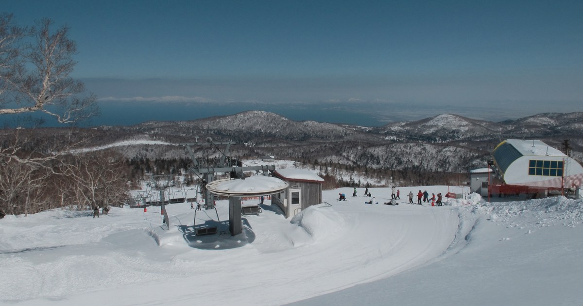 The summit area of Sapporo Kokusai on a clear winter day, a chairlift top station and hut with skiers on the snow, snow-covered ridges behind and a sheet of sea on the horizon