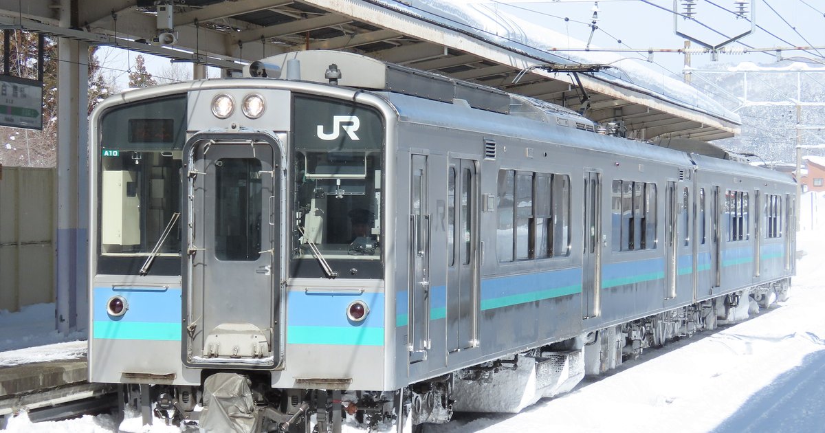 冬天積雪的白馬站月台旁，停著一列車頭有 JR 標誌、銀色車身配藍綠色腰帶的 E127 系普通電車，後方是覆雪的山坡