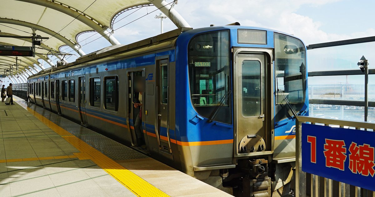 仙台空港站月台上停著一列藍色車頭、銀色車身的仙台空港鐵道 SAT721 系列車，車頭貼著編號 SA103，月台上方是白色拱形帳篷式頂棚，右側欄杆掛著紅字藍底的「1番線」牌，欄杆外看得到機場停車場
