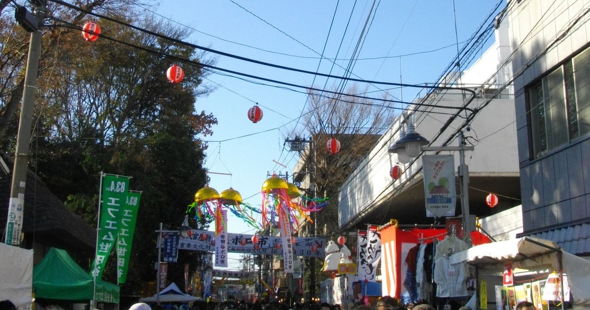 2012 年 12 月晴天的世田谷舊貨市集:ボロ市通り上空掛著紅燈籠與垂著彩帶的金色彩球,兩側是白色與綠色的攤位帳篷、掛著衣服的攤子與綠色旗幟,畫面下緣是擠滿巷道的人群頭頂